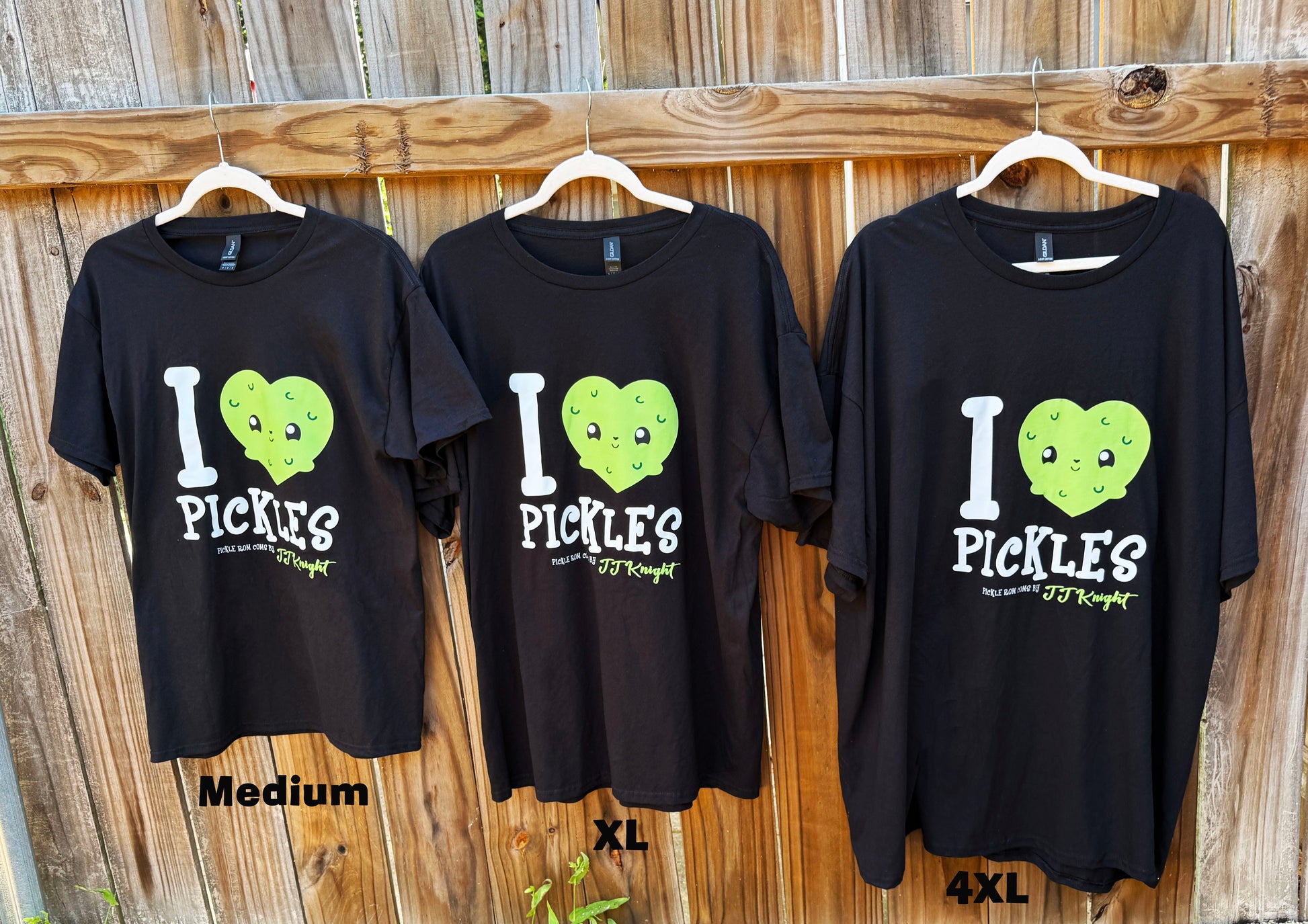 Three black t-shirts with 'I ❤️ Pickles' design on hangers against a wooden fence.
